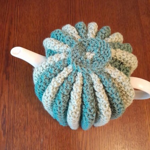 May include: A hand-crocheted tea cosy in teal, grey, and cream. The cosy has a flower-like design on top and vertical ribbed detailing. It is designed to fit over a teapot. The cosy is on a white teapot spout, set against a wooden surface.