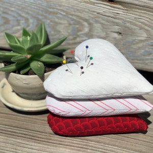 May include: A stack of three heart-shaped pincushions. The top pincushion is white with a quilted texture and has several colorful pins stuck in it. The middle pincushion is red and white striped. The bottom pincushion is red with a scalloped edge.
