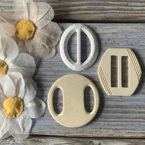 May include: Three vintage white plastic belt buckles. The buckles are oval shaped with a single or double loop. The buckles have a smooth, slightly textured surface.
