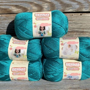 May include: Six teal-colored yarn skeins stacked on a wooden surface. Each skein has a label that reads "BERNAT Softee Baby" with an image of a baby or child. The yarn is a soft, bulky weight, ideal for baby blankets and other projects.