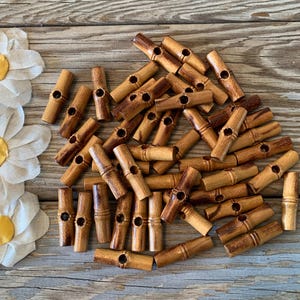 May include: A collection of wooden toggle buttons in various shades of brown, scattered on a weathered wooden surface. Each button has a cylindrical shape with a central hole and a groove for sewing. The buttons are approximately 1 inch long.
