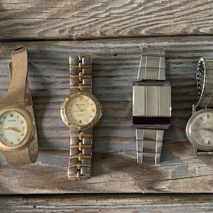 May include: Four vintage wristwatches with different band styles are displayed on a wooden surface. The watches have gold, silver, and two-tone metal bands. The watch faces are white or cream-colored with black numerals and hands. One watch has a mesh band.