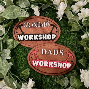 May include: Two wooden signs with the text "Grandad's Workshop" and "Dad's Workshop" in white lettering. The signs are oval shaped and have a brown wood grain finish. Each sign has a pair of black hammer silhouettes on either side of the text.