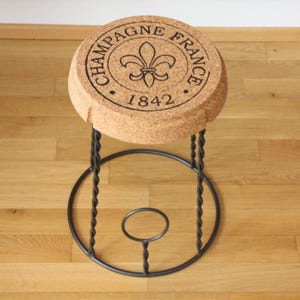 Op de afbeelding: Een kruk met een zitting in de vorm van een grote champagnekurk van lichtbruin kurkmateriaal. De bovenkant is bedrukt met de tekst CHAMPAGNE FRANCE 1842 en een fleur-de-lis symbool. Het heeft een zwart metalen onderstel met gedraaide poten.