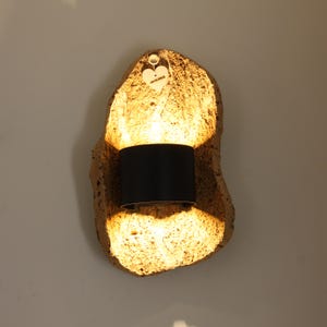 Puede incluir: Una lámpara de pared con una base texturizada de color marrón dorado. La lámpara presenta una pantalla cilíndrica negra y emite un brillo cálido. Una pequeña etiqueta de madera en forma de corazón cuelga encima.