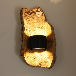 Puede incluir: Una lámpara de pared con una parte trasera de piedra natural texturizada de color marrón claro. Una pantalla de lámpara cilíndrica negra está centrada en la piedra, emitiendo un brillo cálido. Una pequeña etiqueta de madera en forma de corazón cuelga.