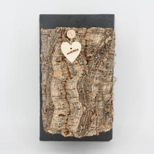Puede incluir: Un trozo rectangular de corteza de corcho natural montado sobre una pizarra negra. Un pequeño corazón de madera con el texto "verkORKst" está sujeto con hilo. El corcho tiene un aspecto texturizado, marrón y beige.