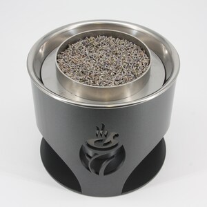 May include: A dark gray aromatherapy diffuser with a stainless steel interior and a black base. The diffuser holds dried lavender buds. A decorative cut-out design of a stylized flame is on the side.