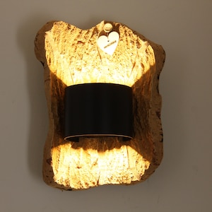 Puede incluir: Una lámpara de pared con una base natural y texturizada de color marrón claro. La lámpara presenta una pantalla cilíndrica negra. Un pequeño corazón de madera con la palabra "verliebt" está adjunto en la parte superior.