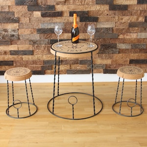 Puede incluir: Un conjunto de tres muebles de metal negro con tapas de corcho. La mesa y los dos taburetes presentan una tapa de corcho circular con un logotipo en blanco y negro que dice "Champagne Perrier Jouet 1842".