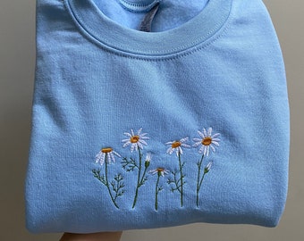 May include: A light blue sweatshirt with embroidered white daisies and yellow centers. The daisies are arranged in a row across the front of the folded garment. The sweatshirt has a crew neck and long sleeves.
