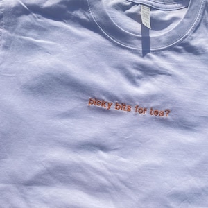 May include: A white t-shirt with the embroidered text "picky bits for tea?" in orange. The shirt is made of a soft material and has a crew neck. The image is taken outdoors, with grass visible in the background.
