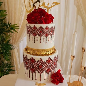 May include: A two-tier wedding cake decorated with red and gold accents. The cake is adorned with intricate embroidery-like patterns and a gold band with coins. The top tier features a red flower arrangement and a gold topper with Arabic calligraphy.