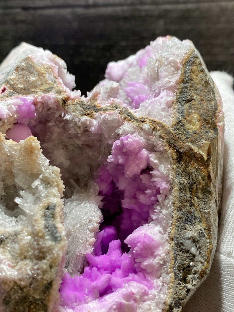 May include: A geode with a cavity filled with vibrant purple crystals. The outer rock is a mix of grey, brown, and white, with a rough texture. The crystals inside are a deep, saturated purple, creating a striking contrast.