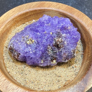 Puede incluir: Un c&uacute;mulo de cristales de amatista p&uacute;rpura con una superficie rugosa y texturizada. El cristal est&aacute; sentado en un cuenco de madera lleno de arena.
