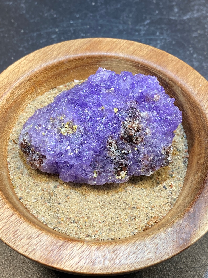 Puede incluir: Un c&uacute;mulo de cristales de amatista p&uacute;rpura con una superficie rugosa y texturizada. El cristal est&aacute; sentado en un cuenco de madera lleno de arena.