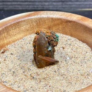 May include: A brown and green crystal with a clear quartz point, sitting in a wooden bowl filled with sand.