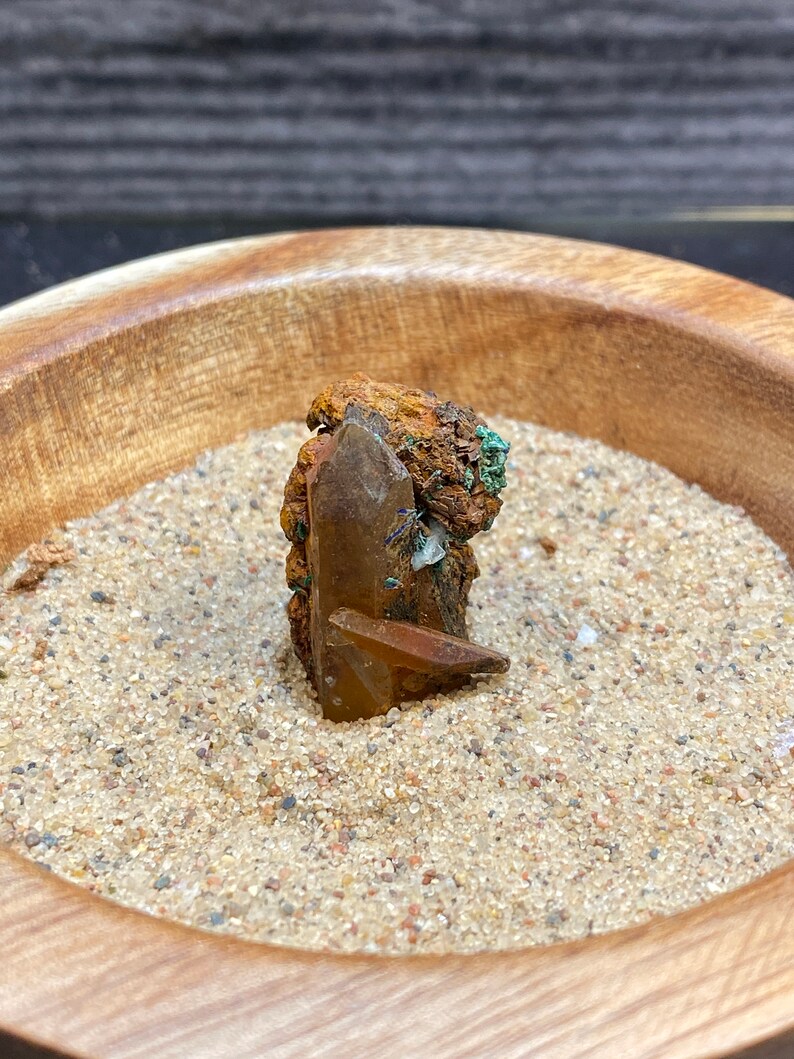 May include: A brown and green crystal with a clear quartz point, sitting in a wooden bowl filled with sand.