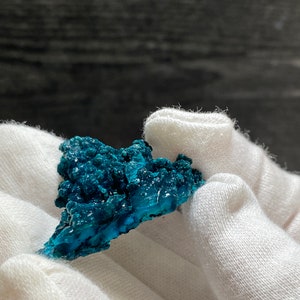 May include: A teal blue, rough-textured, mineral specimen with a jagged edge. The mineral is held in a white, fabric-covered hand.