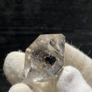 May include: A clear, faceted, octahedral crystal with a dark gray inclusion. The crystal is held in a white, fabric-covered hand.