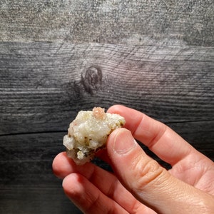May include: A small cluster of white crystals with a pink center, held in a hand against a wood background.