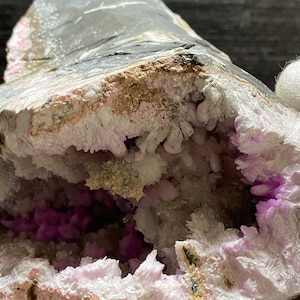 May include: A close-up of a geode. The exterior is grey, and the cavity is filled with white and purple crystals. The geode's outer layer has shades of grey and pink, while the interior crystals sparkle.