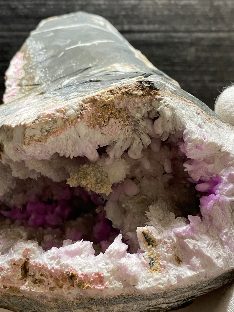 May include: A close-up of a geode. The exterior is grey, and the cavity is filled with white and purple crystals. The geode's outer layer has shades of grey and pink, while the interior crystals sparkle.