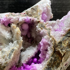 May include: A close-up of a mineral specimen with a cavity filled with vivid purple crystals. The outer matrix is a mix of white, grey, and brown, creating a striking contrast. The crystals have a sparkling, textured appearance, showcasing natural formations.