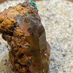 May include: A large, clear quartz crystal with a brown, rusty coating. The crystal is surrounded by smaller, brown, rusty crystals.