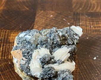 May include: A cluster of white barite crystals on a matrix of gray and black minerals. The barite crystals are translucent and have a pearly luster. The matrix is rough and textured.