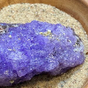 Puede incluir: Primer plano de un cristal de amatista p&uacute;rpura con una superficie rugosa y texturizada. El cristal est&aacute; sentado sobre un lecho de arena fina.