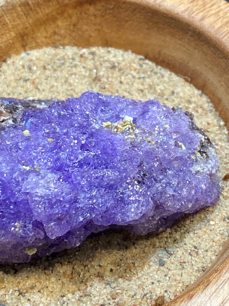 Puede incluir: Primer plano de un cristal de amatista p&uacute;rpura con una superficie rugosa y texturizada. El cristal est&aacute; sentado sobre un lecho de arena fina.