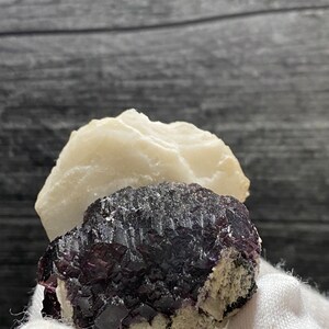 Könnte beinhalten: Ein Cluster aus violetten Fluoritkristallen mit einer weißen Calcitmatrix. Die Fluoritkristalle sind dunkelviolett und haben einen glasartigen Glanz. Die Calcitmatrix ist weiß und hat eine raue Textur.