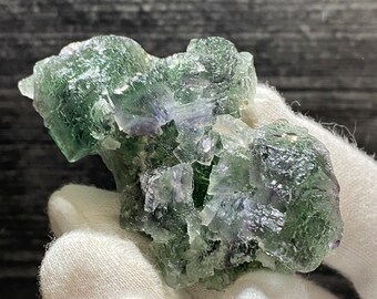 Könnte beinhalten: Ein Cluster aus grünen Fluoritkristallen mit violetten und weißen Einschlüssen. Die Kristalle sind in einem zufälligen Muster angeordnet und haben eine raue, natürliche Textur.