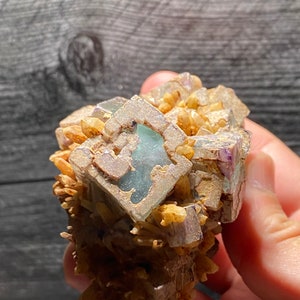 以下が含まれることがあります： 天然の蛍石の結晶のクラスター。結晶は、紫、緑、茶色など、さまざまな色を示しています。蛍石は、小さな金茶色の水晶の結晶を背景に配置されています。
