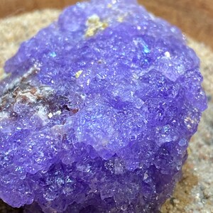 Puede incluir: Primer plano de un c&uacute;mulo de cristal de amatista p&uacute;rpura con una superficie rugosa y texturizada. El cristal est&aacute; rodeado de arena.