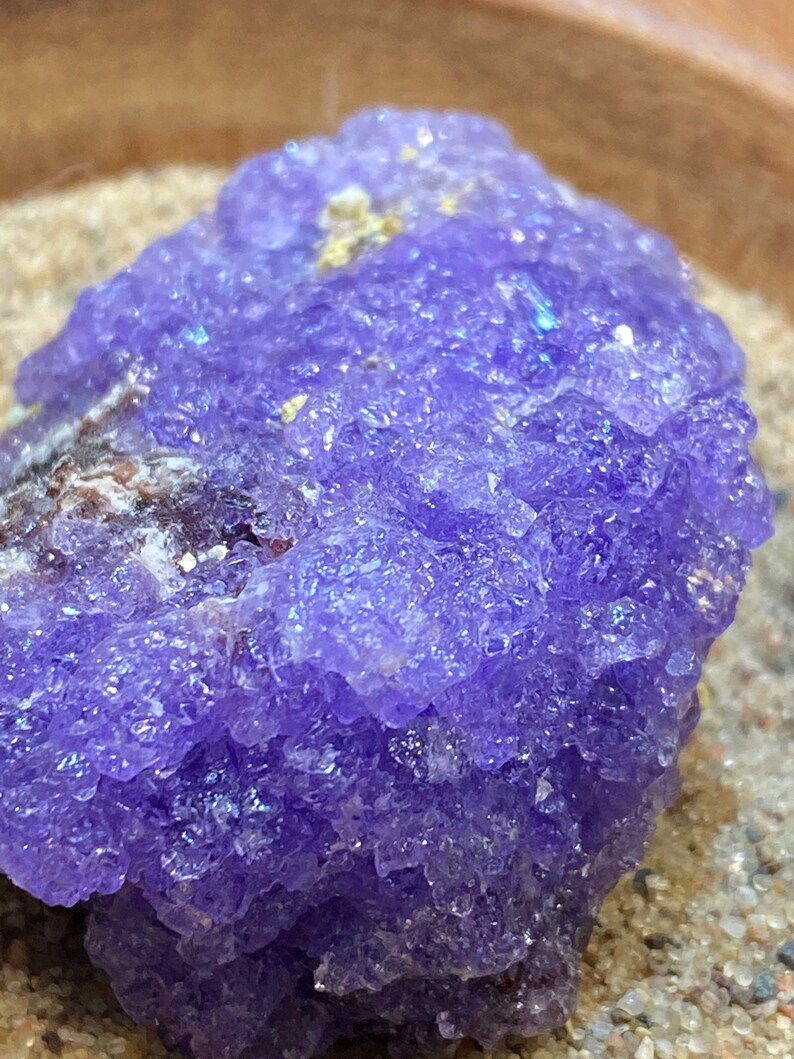 Puede incluir: Primer plano de un c&uacute;mulo de cristal de amatista p&uacute;rpura con una superficie rugosa y texturizada. El cristal est&aacute; rodeado de arena.