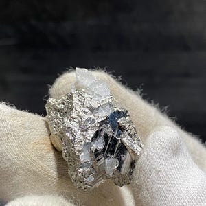 May include: A close-up of a pyrite specimen with a quartz crystal. The pyrite has a metallic silver color and a rough, textured surface. The quartz crystal is clear and sits atop the pyrite. The specimen is held in a white glove.
