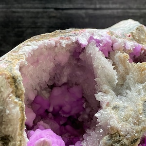 May include: Close-up of a geode featuring a cavity filled with vivid purple crystalline structures. The outer shell displays beige and grey hues, with white and pink details. The inner crystals have a clustered, rounded form.