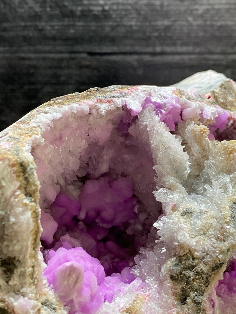 May include: Close-up of a geode featuring a cavity filled with vivid purple crystalline structures. The outer shell displays beige and grey hues, with white and pink details. The inner crystals have a clustered, rounded form.