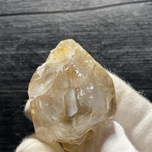 May include: A clear, faceted, golden citrine crystal with a pointed tip. The crystal is held in a white gloved hand.