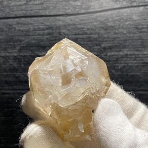 以下が含まれることがあります： 先のとがった頂上を持つ、透明で面取りされた薄い茶色のクリスタル。クリスタルは白い布で覆われた手で持たれています。
