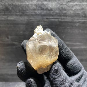 以下が含まれることがあります： 黒い手袋に収められた、金色の色合いの透明なファセットクリスタル。クリスタルは複雑な幾何学的形状をしており、ざらざらした質感の基部を持っています。光がファセットを捉え、きらめく効果を生み出しています。天然の鉱物標本です。