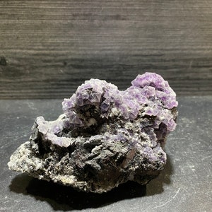 May include: A cluster of purple fluorite crystals on a dark gray matrix. The crystals are sharp and well-formed, with a glassy luster.
