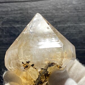May include: A clear quartz crystal with a pointed, pyramidal shape. The crystal has a light yellow hue and some dark inclusions near the base. The crystal is held by a gloved hand. The crystal is a natural mineral specimen.
