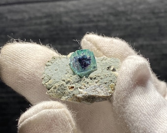 May include: A small, green and blue fluorite crystal on a piece of rock. The crystal is in the shape of a cube and has a glassy luster.