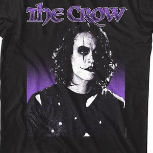May include: Black t-shirt featuring a portrait of a person with white face makeup and dark eye makeup, with the text "The Crow" in purple above the portrait. The background is purple.