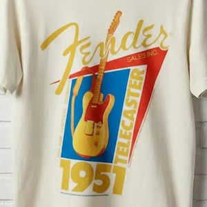 May include: Cream-colored t-shirt featuring a Fender Telecaster guitar graphic. The design includes the Fender logo in gold script, a red and blue triangle, and the text "TELECASTER 1951". The shirt is hanging on a gray hanger against a white brick wall.