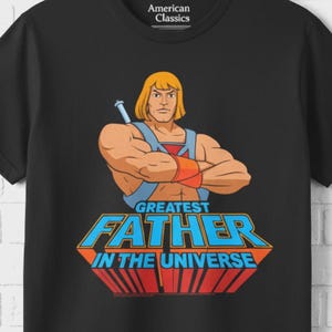 May include: Black t-shirt featuring a cartoon illustration of a muscular blond man with a sword, and the text "GREATEST FATHER IN THE UNIVERSE" in blue and red. The shirt has a crew neck and the brand "American Classics" is printed on the upper left.