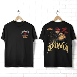 May include: Two black t-shirts displayed on hangers. The front features a small Akuma logo and Street Fighter text. The back displays a large graphic of the character Akuma with red hair, the red kanji symbol, and the yellow text Akuma.